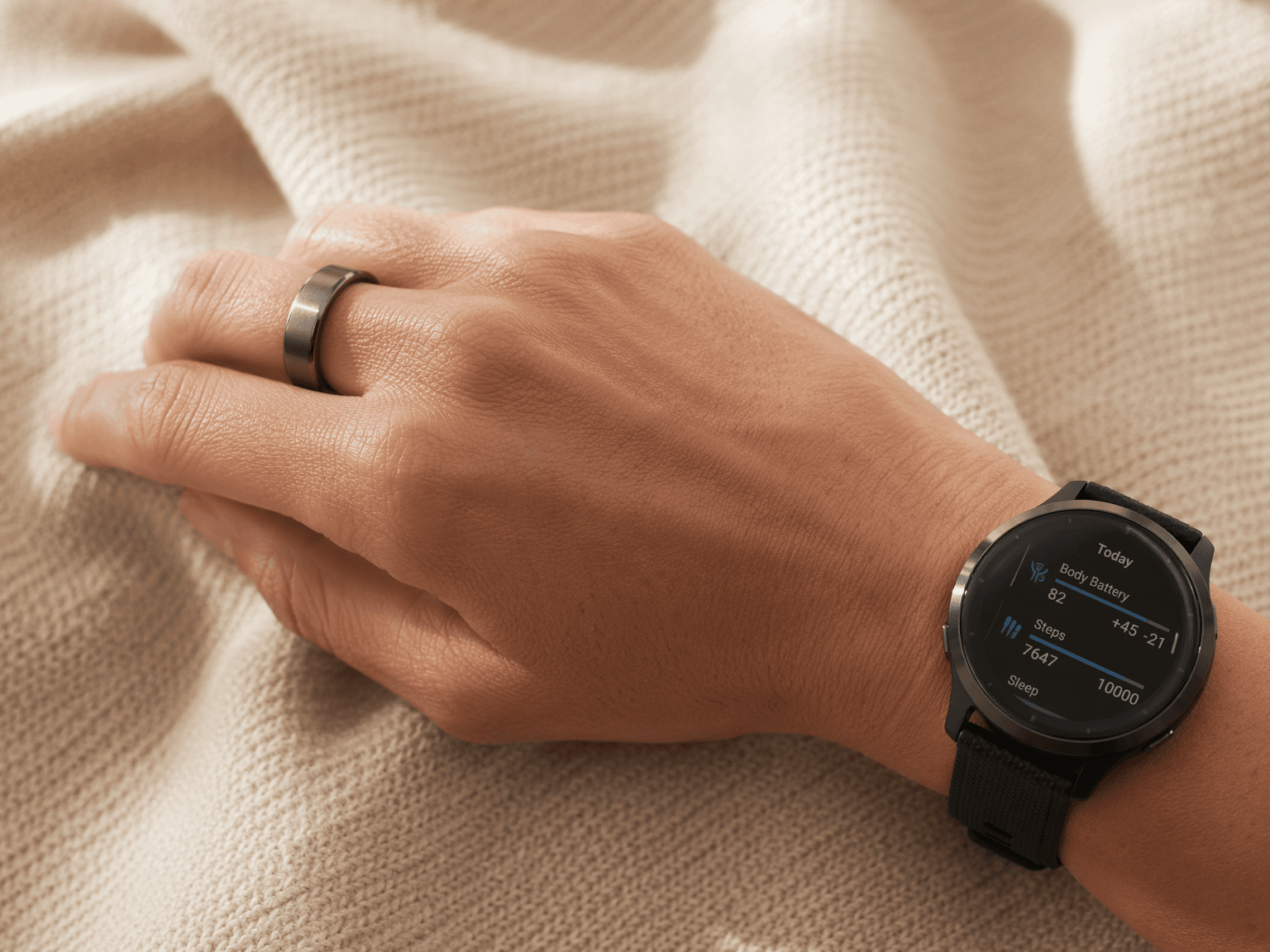 Close-up of a hand and wrist wearing a silver smart ring on the index finger and a round smartwatch on the wrist, on a warm neutral textile background in soft morning light.