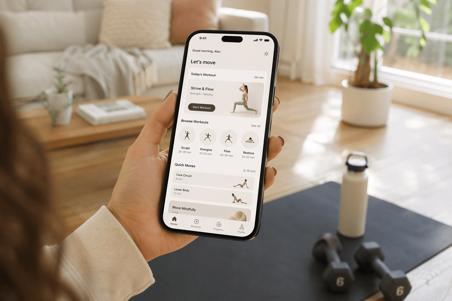 A smartphone held in a woman's hand in a sunlit living room with a yoga mat, water bottle, and small dumbbells on the floor; the phone screen displays a fitness app workout interface against warm neutral-toned home decor with soft window light.