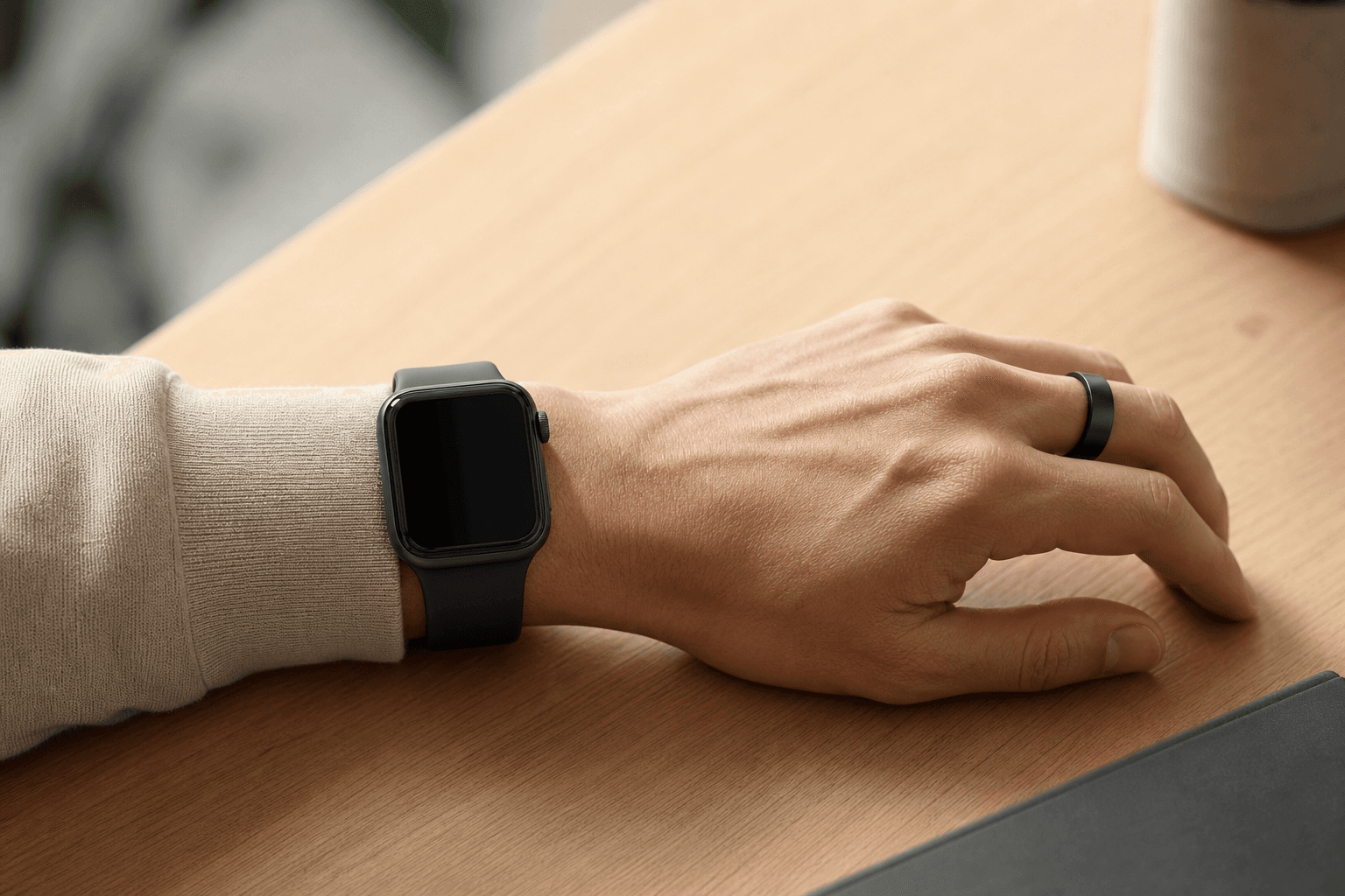 A person's forearm, wrist, and hand resting on a wooden desk, with a smartwatch on the wrist and a smart ring on the index finger of the same hand, illustrating a hybrid wearable setup.