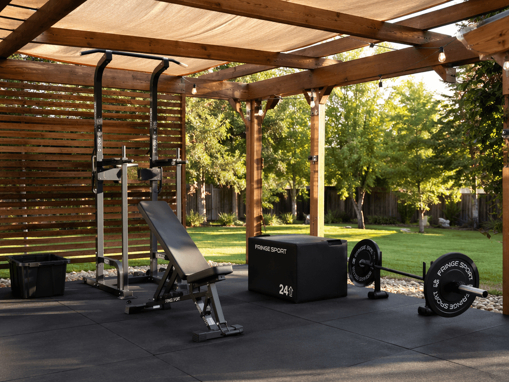 Organized backyard home gym setup under a wooden pergola with power tower, adjustable bench, plyo box, and barbell on rubber mats.