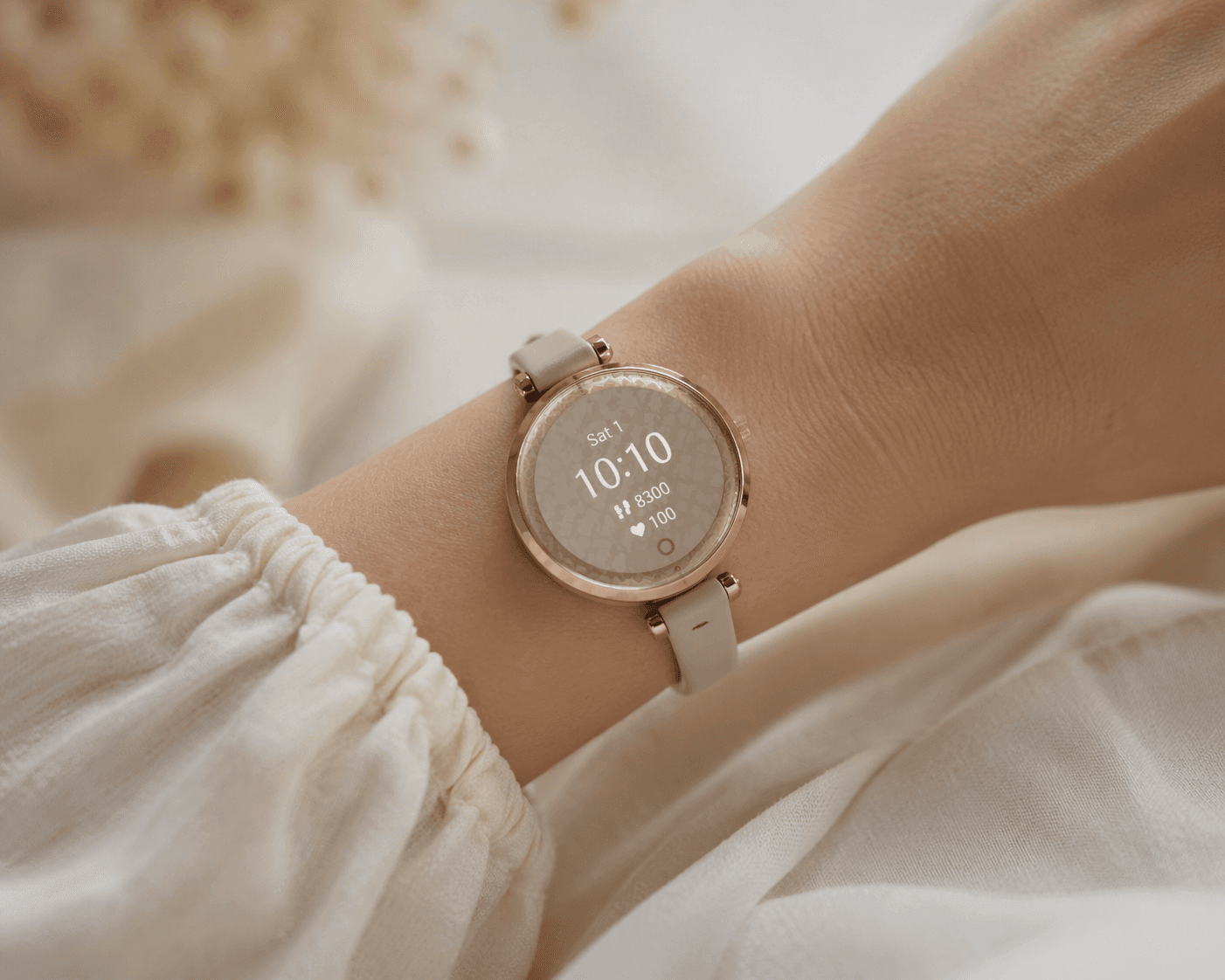 A small round jewelry-inspired fitness smartwatch with an ornate patterned dial worn on a very petite woman's wrist, with the band fitting snugly and proportionally. Soft linen sleeve visible in the background.