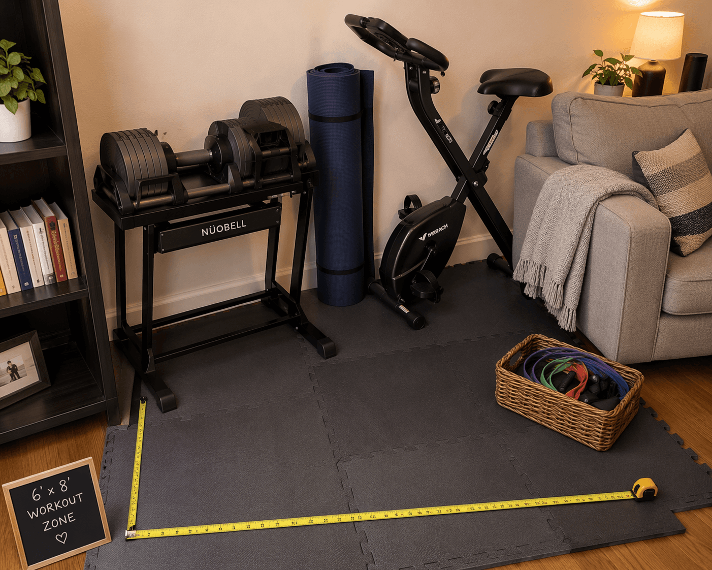 A warm residential flat-lay photograph of a compact home gym in an apartment corner showing adjustable dumbbells on a stand, a folded yoga mat, a small folding exercise bike beside a couch, a basket of resistance bands, and interlocking foam tiles, with a measuring tape on the floor indicating the space dimensions.