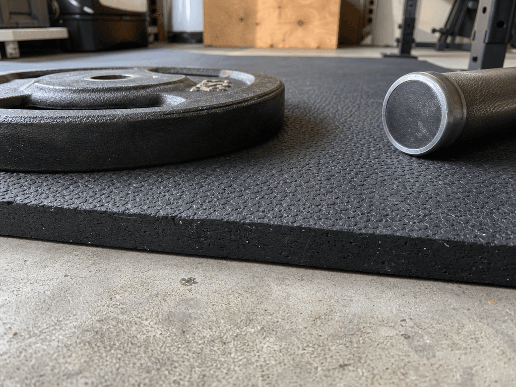 A close-up low-angle view showing the edge profile of a 3/4-inch thick black rubber horse stall mat on a concrete garage floor, with a cast iron weight plate and a barbell end cap resting on the mat surface.