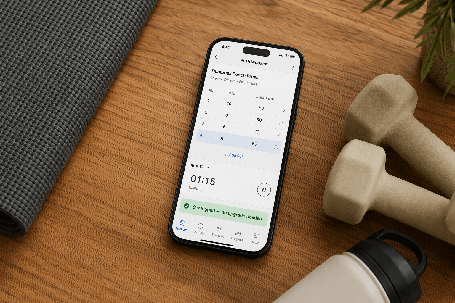Flat-lay photo on a wooden desk of a smartphone showing a workout logging interface with a notification 'Set logged — no upgrade needed,' beside a yoga mat, light dumbbells, and a water bottle.