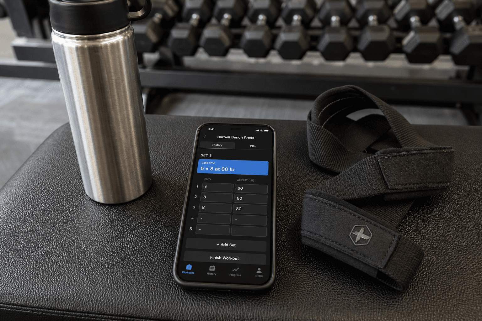 A flat-lay composition on a gym bench: a smartphone centered with the screen showing a workout tracker interface displaying 'last time: 5×8 at 80 lb' in a highlighted row. Beside the phone are a metal water bottle and black lifting straps. In the softly blurred background, a dumbbell rack is visible.