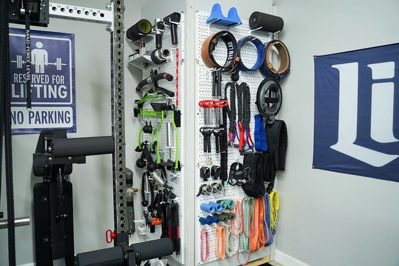 A Wall Control metal pegboard mounted on a garage wall, holding organized gym accessories including resistance bands, jump ropes, and small equipment on hooks and attachments.