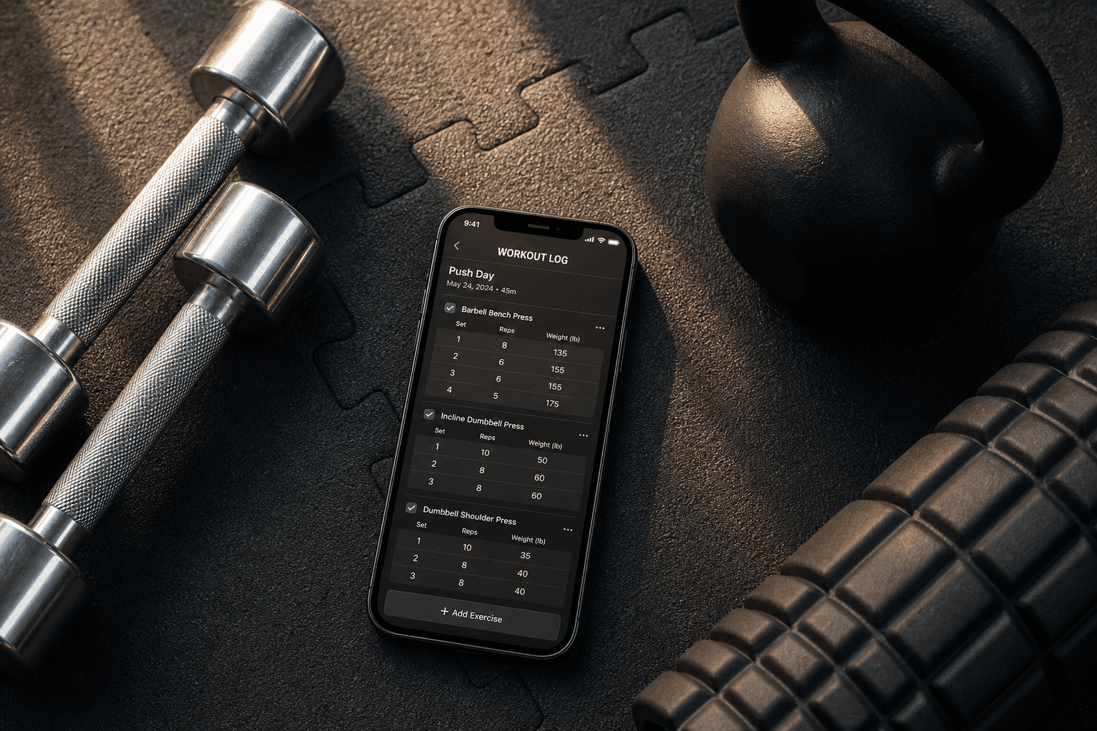 Top-down view of a smartphone on a dark gym mat showing a workout log with sets and weights, surrounded by dumbbells, a kettlebell, and a foam roller under warm natural light.