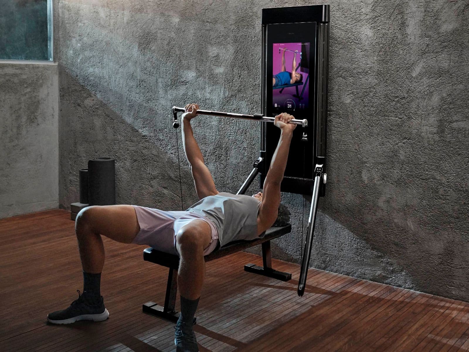Person bench pressing on a Tonal 2 smart home gym with digital resistance arms and screen visible