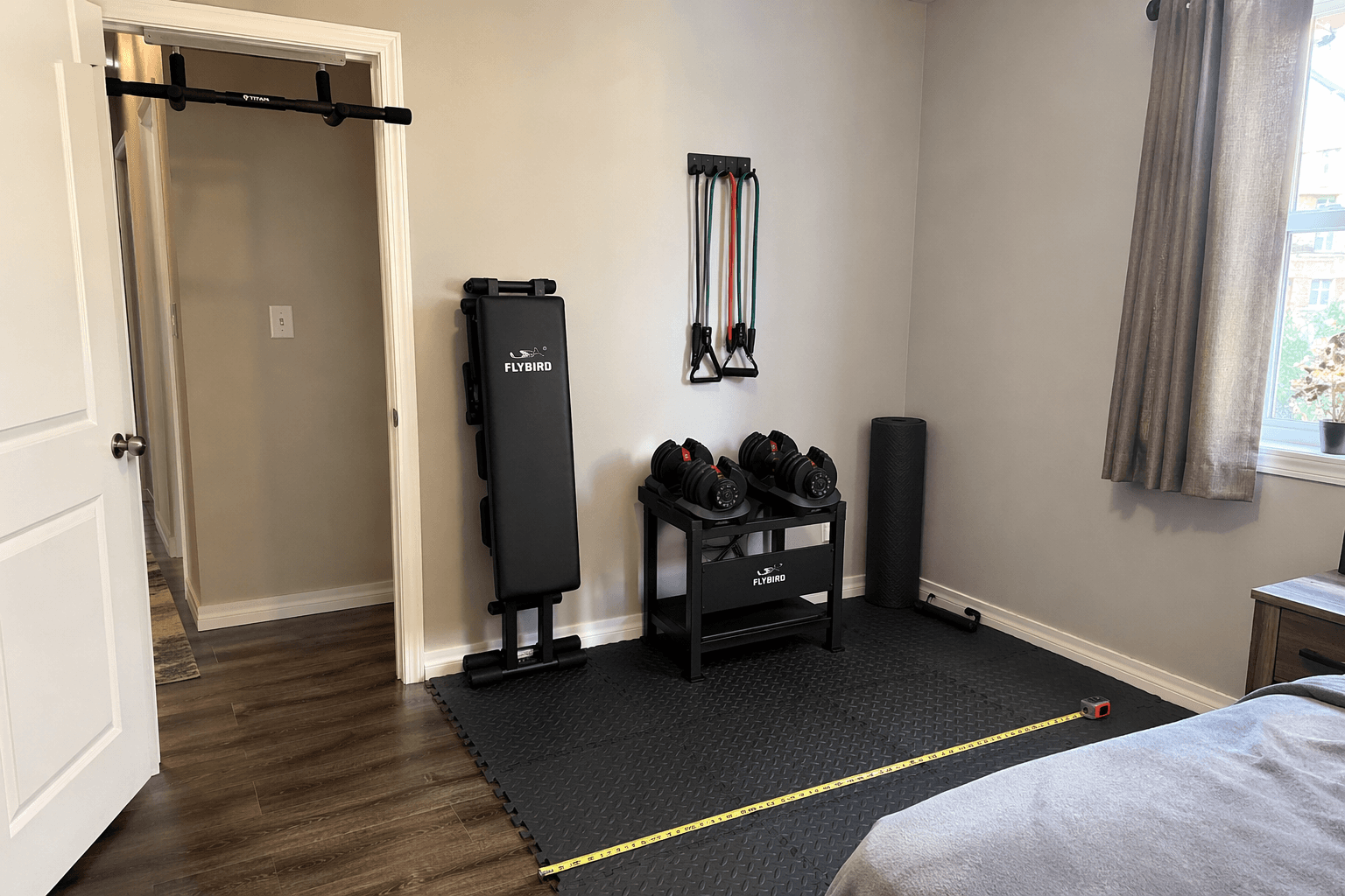 A realistic compact home gym setup in a small apartment bedroom corner with adjustable dumbbells on a stand, an adjustable bench folded and stored upright, a doorway pull-up bar on the door frame, a rolled yoga mat, resistance bands on a wall hook, interlocking foam floor tiles, and a tape measure visible on the floor showing dimensions.