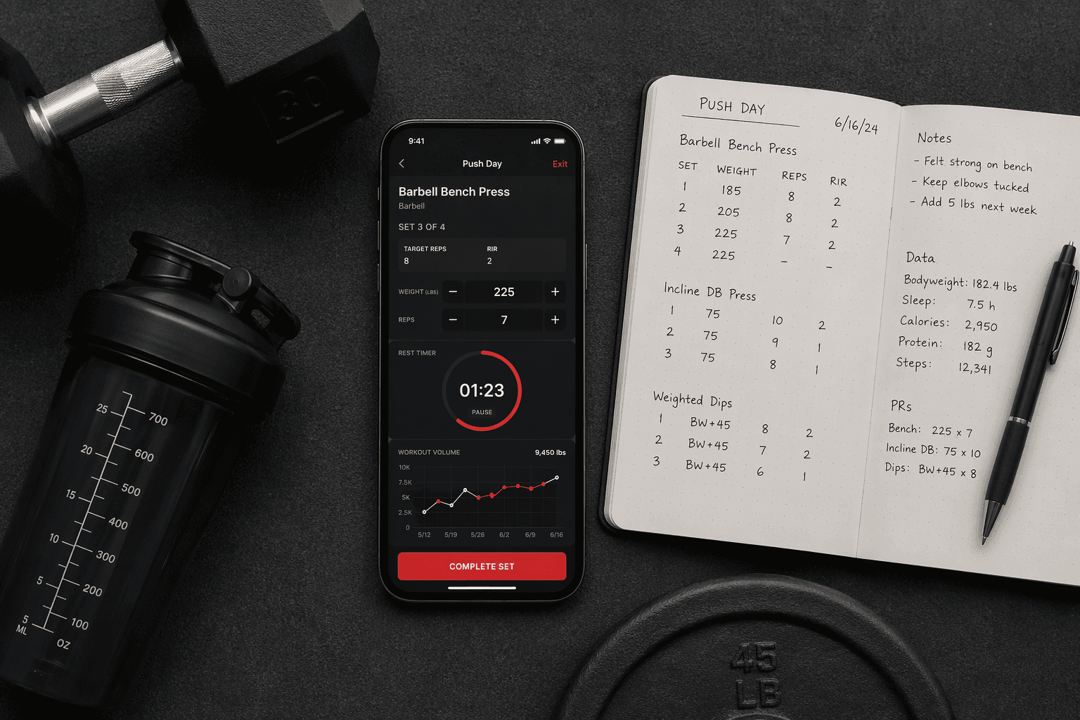 Flat-lay composition on a dark grey gym mat showing a smartphone with a workout tracker app mid-session displaying exercise name, set/rep tracking, rest timer, and a progress chart, alongside an open notebook with handwritten training logs and a pen.