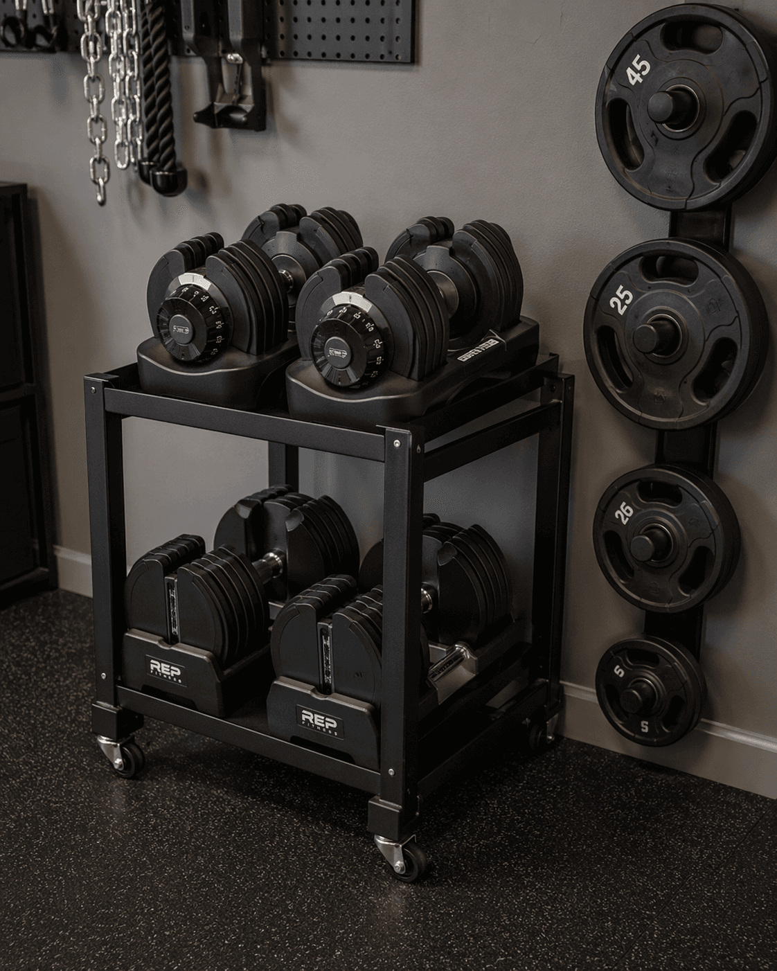 Adjustable dumbbells on a compact black metal storage cart in a garage gym, with a vertical plate tree mounted on the wall behind them.