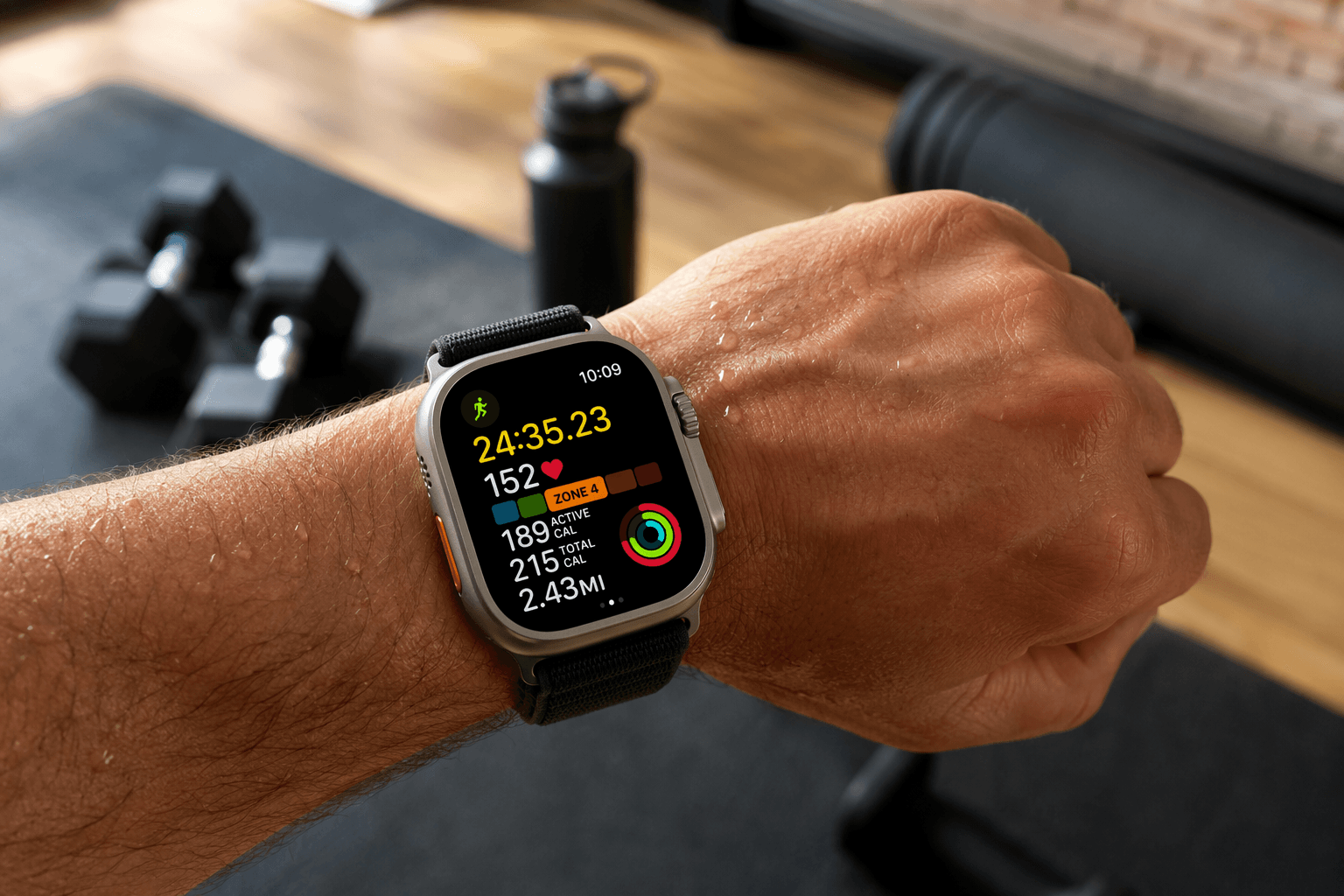 A close-up of a person's wrist wearing an Apple Watch Ultra 3 during an active home workout. The watch display shows real-time workout metrics including activity rings, heart rate data, and a workout timer. In the softly blurred background, home fitness equipment such as rubber dumbbells and a yoga mat are visible.