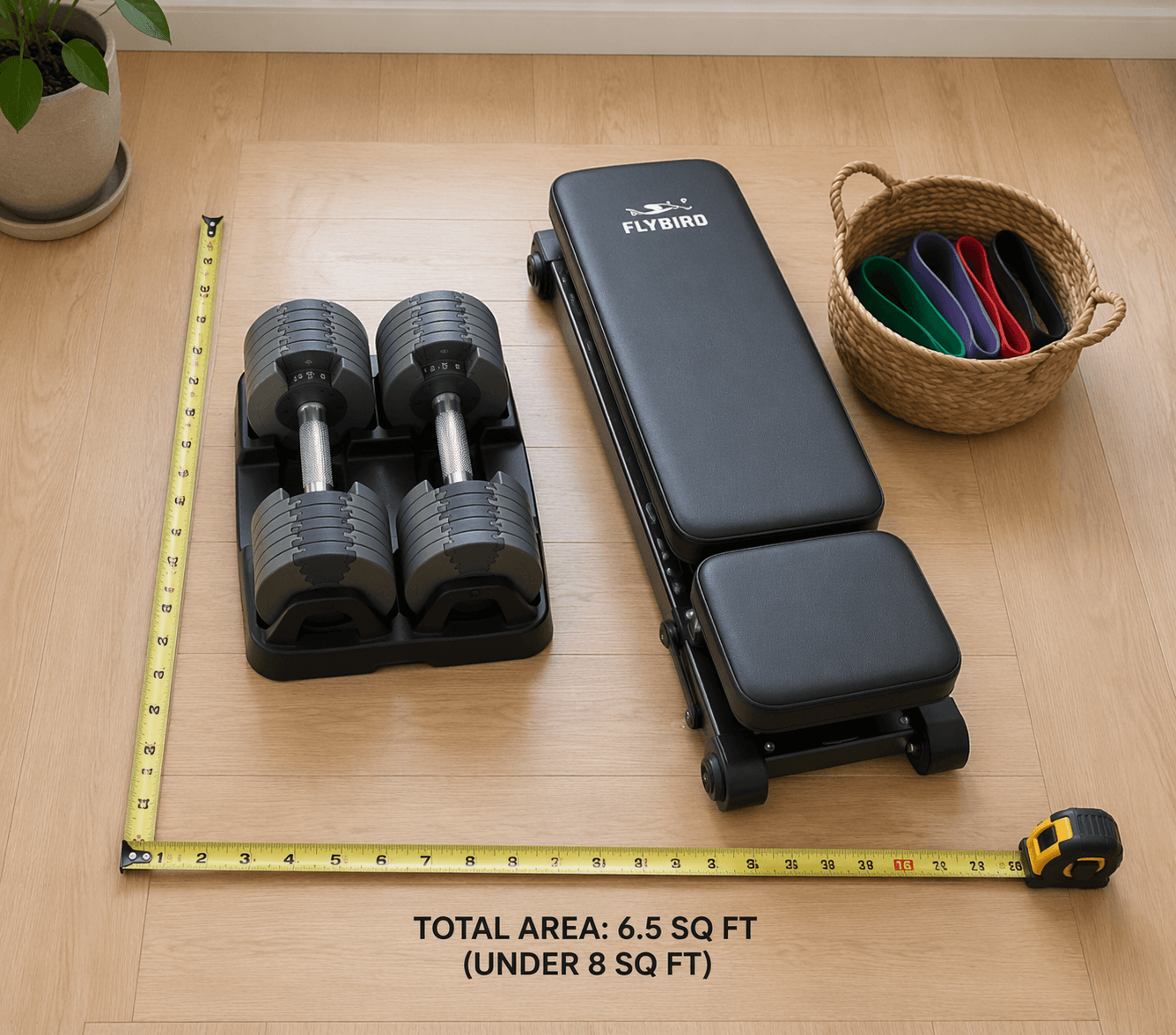 A clean flat-lay photograph of a modular home gym setup: grey adjustable dumbbells in a black cradle beside a compact foldable weight bench, with a small basket of colorful resistance bands. A measuring tape on the light wood floor shows the total area.