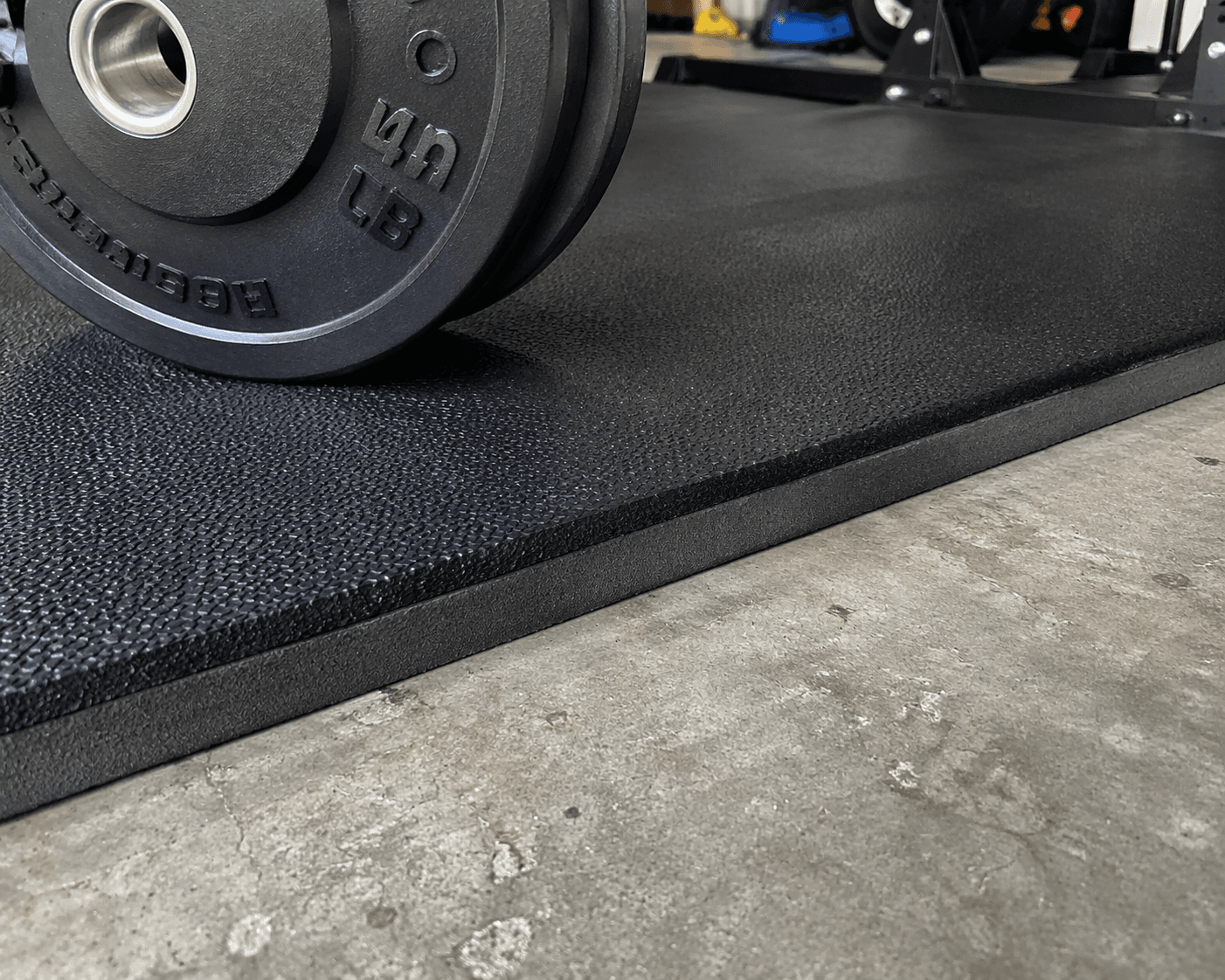 Close-up of the edge where a thick black textured rubber stall mat meets bare gray concrete garage floor. A heavy barbell weight plate rests on the mat surface, demonstrating the thickness and impact protection of the 3/4-inch rubber material over the concrete.