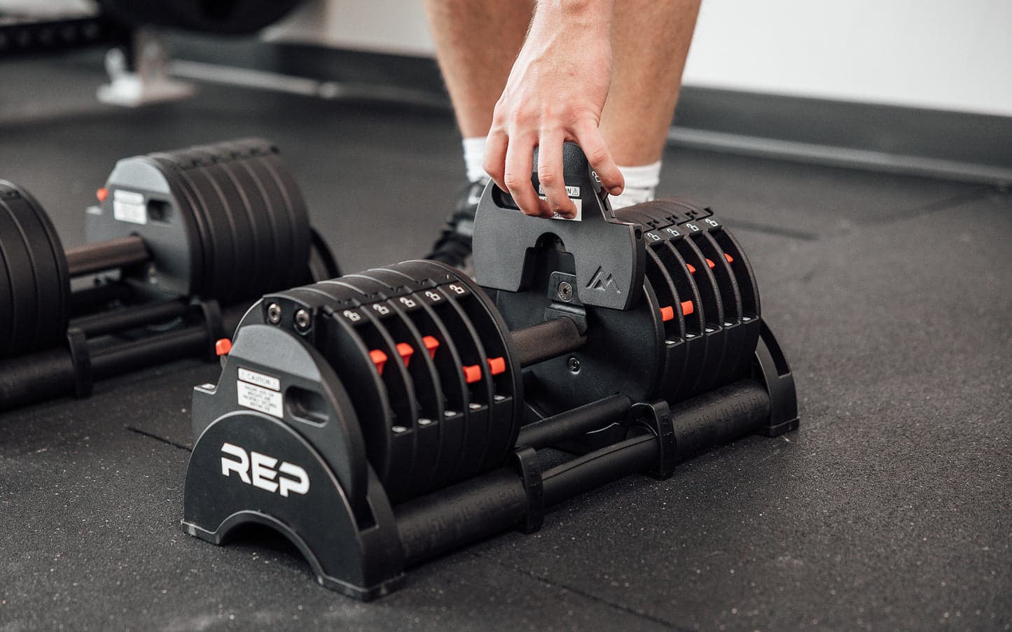 A pair of REP Fitness QuickDraw adjustable dumbbells resting on a home gym floor in a well-lit interior setting, showing the compact space-saving design with the weight adjustment mechanism visible on the side of each dumbbell handle.