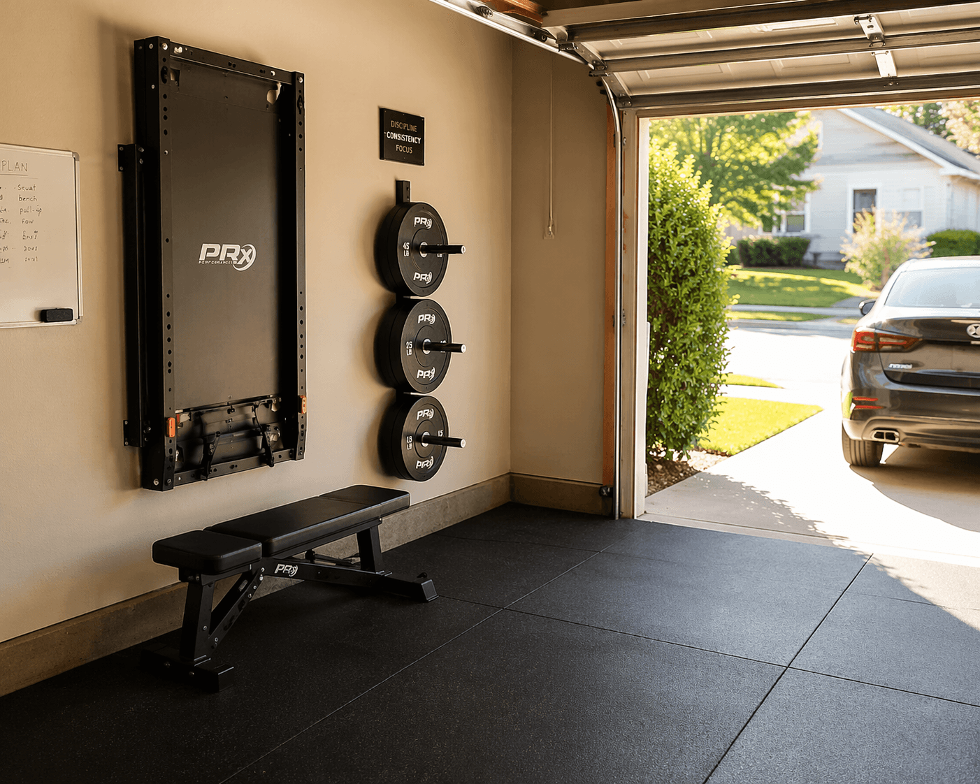 Wide-angle view of a single-car garage gym with a black wall-mounted folding power rack folded flush against the wall, adjustable bench below it, weight plates stored vertically on a wall-mounted tree, dark rubber stall mat flooring, and a clear open center lane.