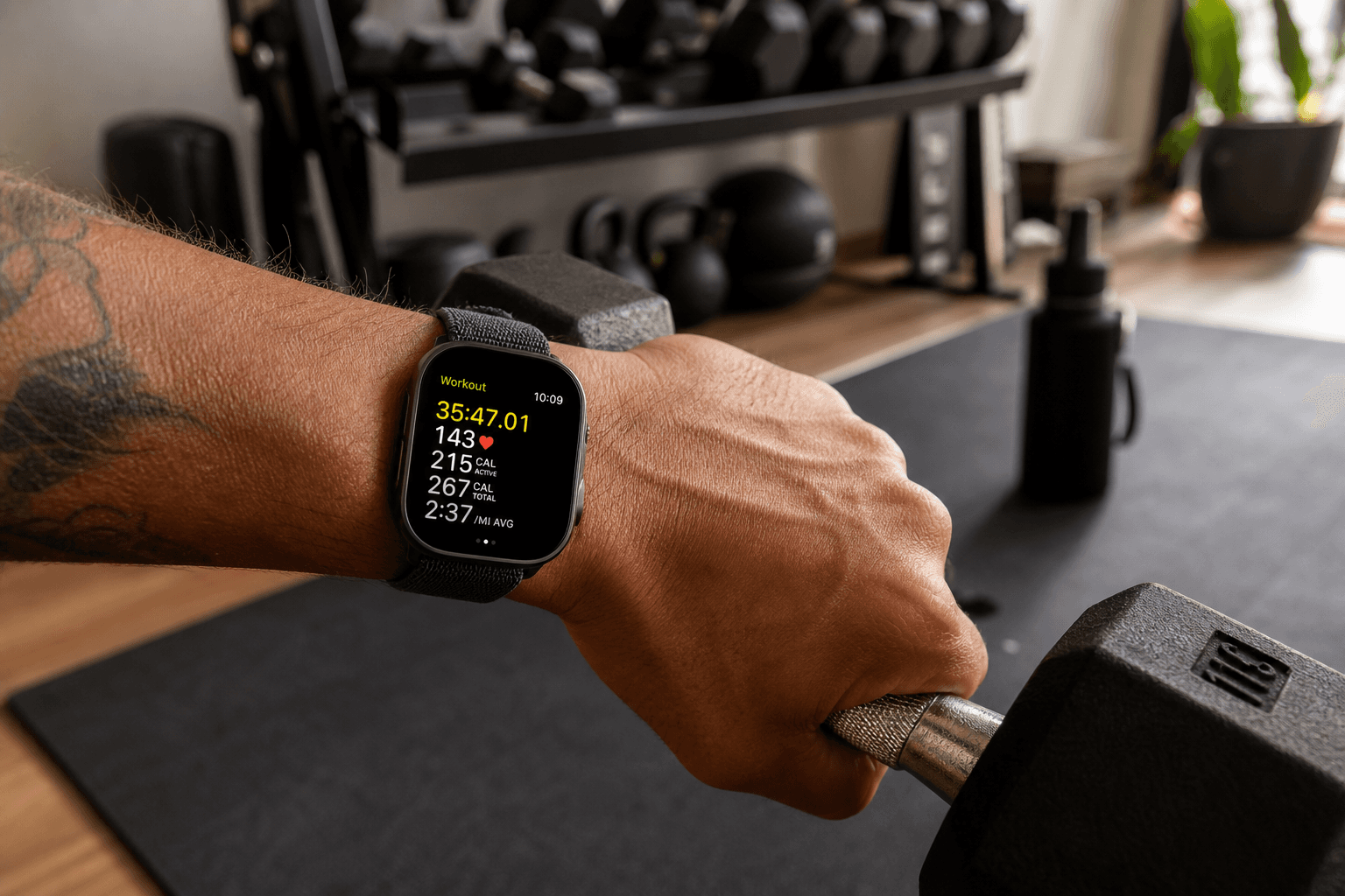 Close-up of a person's wrist wearing a modern fitness smartwatch while gripping a dumbbell in a home gym, with workout equipment visible in the background