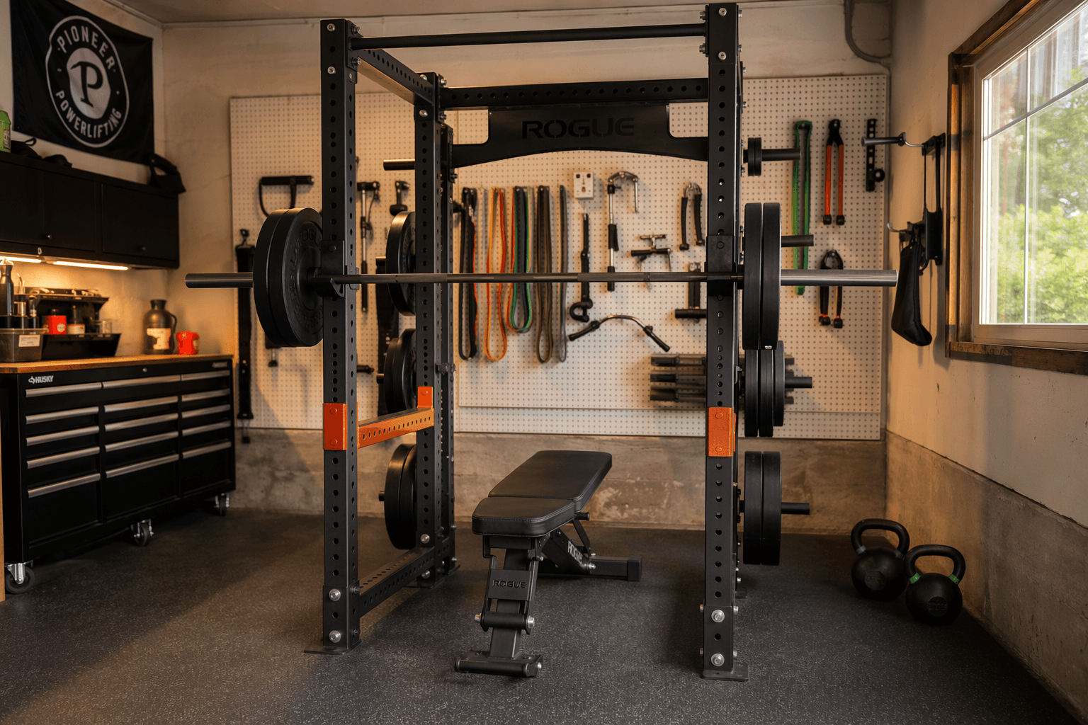 A realistic home garage gym interior with a four-post power rack as the centerpiece, featuring a loaded barbell on J-hooks, orange safety bars, an adjustable bench inside the cage, weight plates stored on rear posts, rubber flooring, pegboard wall with tools, and warm ambient natural light from a side window.