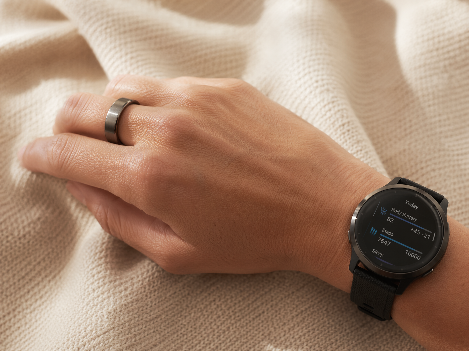 Close-up of a hand and wrist wearing a silver smart ring on the index finger and a round smartwatch on the wrist, on a warm neutral textile background in soft morning light.