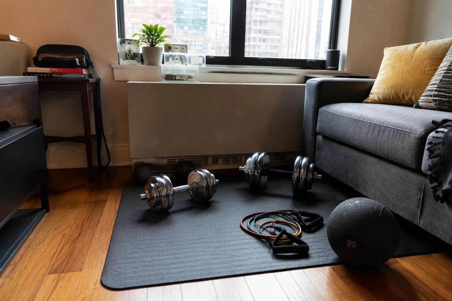 Small apartment living room corner with a rubber gym mat, adjustable dumbbells, resistance bands, and a slam ball beside a sofa