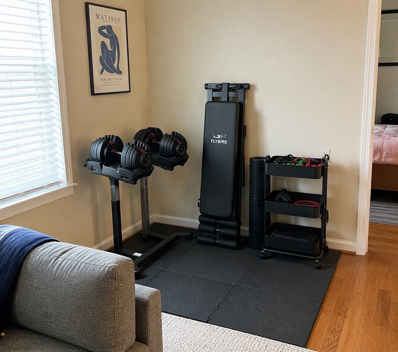 A 6x6 ft apartment gym corner with rubber tiles, adjustable dumbbells, a folded bench, and a rolling storage cart