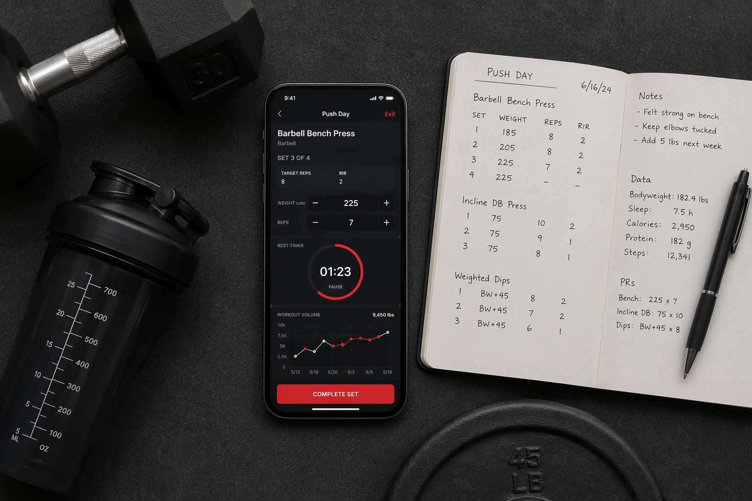 Flat-lay composition on a dark grey gym mat showing a smartphone with a workout tracker app mid-session displaying exercise name, set/rep tracking, rest timer, and a progress chart, alongside an open notebook with handwritten training logs and a pen.