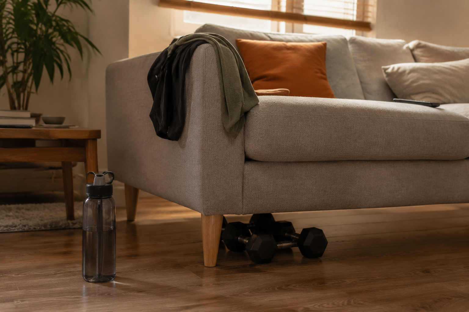 Dumbbells tucked under a sofa in a living room, with a water bottle and workout top nearby