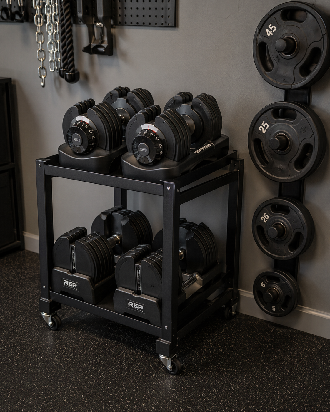 Adjustable dumbbells on a compact black metal storage cart in a garage gym, with a vertical plate tree mounted on the wall behind them.