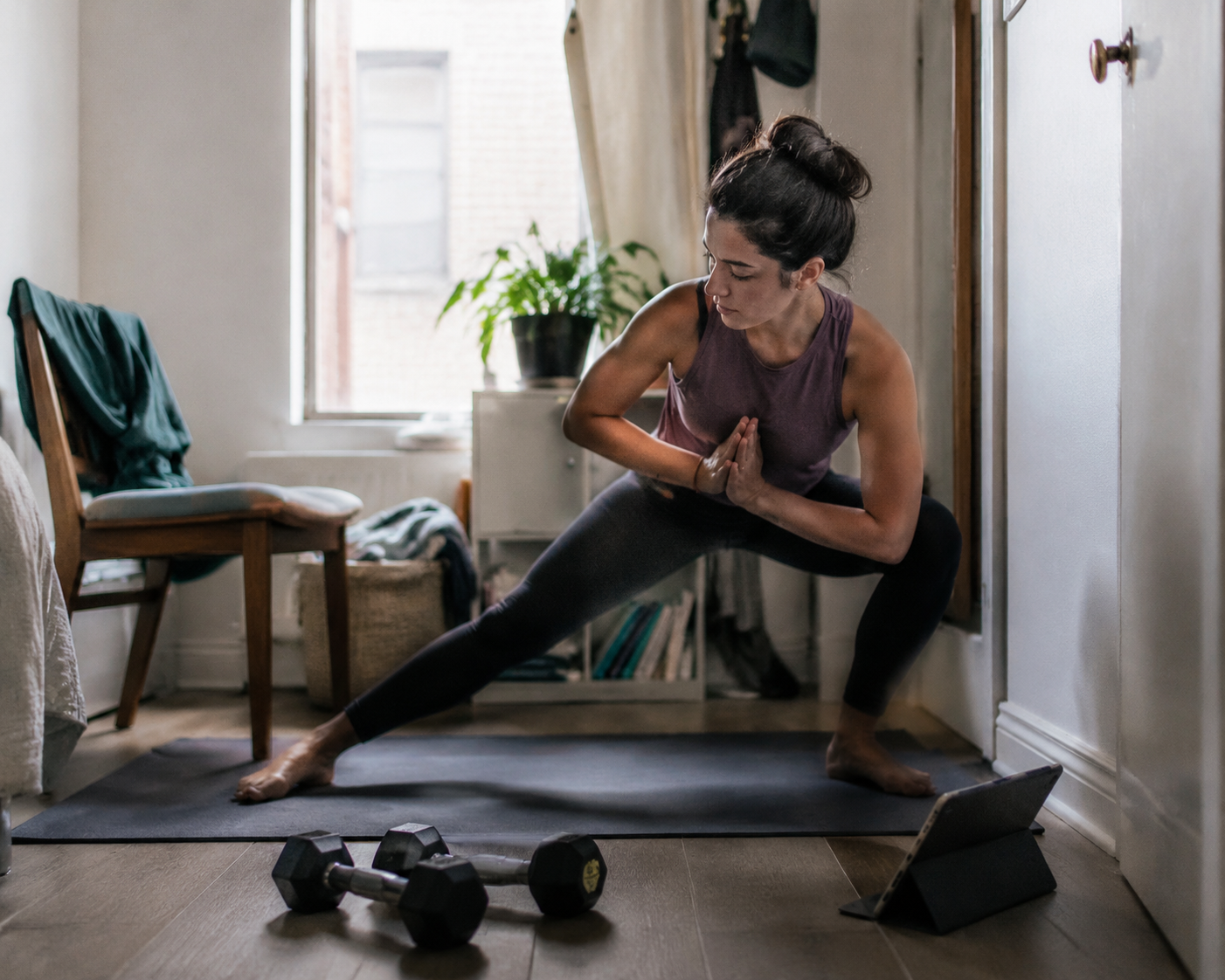 Small apartment workout corner with a yoga mat, adjustable dumbbells, a tablet, and a chair pushed aside