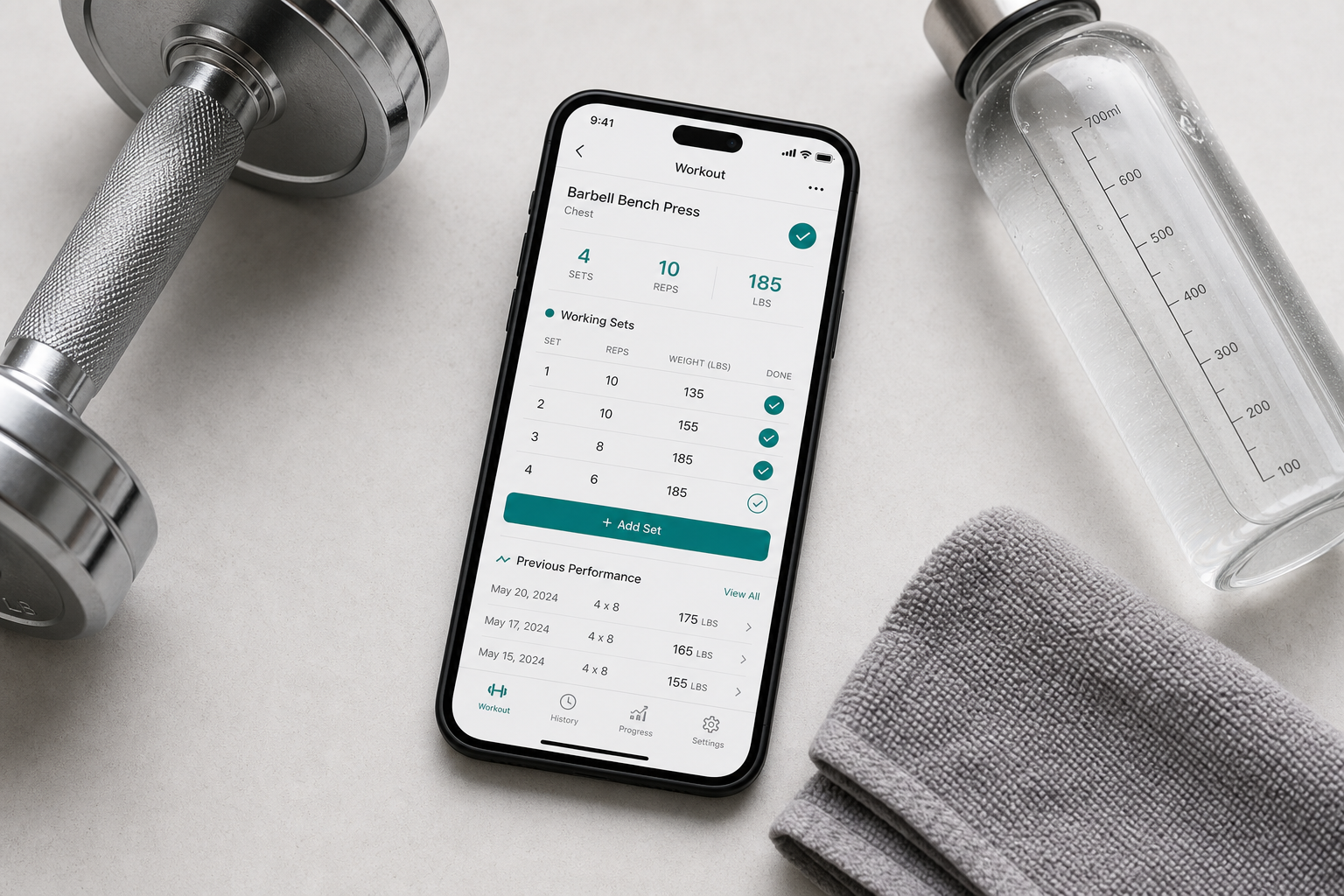 A flat-lay composition of a smartphone on a light surface showing a workout tracking app interface with sets, reps, and weight data, surrounded by a dumbbell, water bottle, and gym towel in muted grays with teal accents