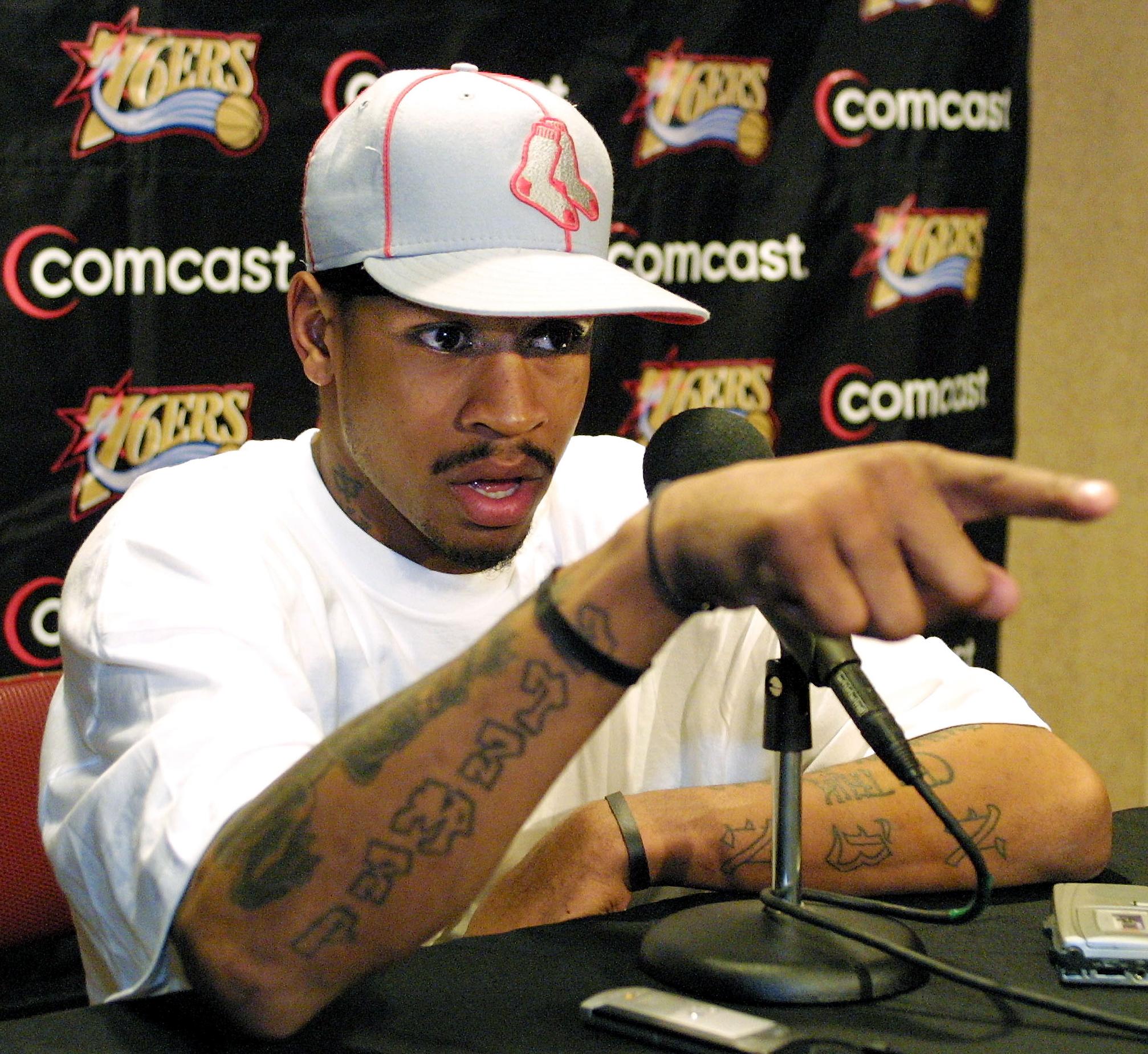 Allen Iverson speaking at a 2002 press conference podium in a Philadelphia 76ers practice jersey and white headband