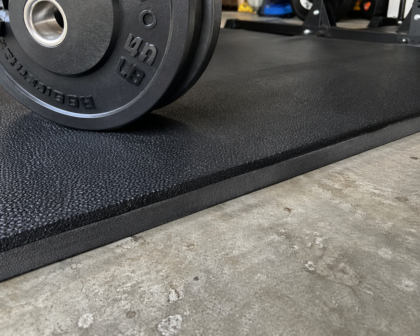 Close-up of the edge where a thick black textured rubber stall mat meets bare gray concrete garage floor. A heavy barbell weight plate rests on the mat surface, demonstrating the thickness and impact protection of the 3/4-inch rubber material over the concrete.