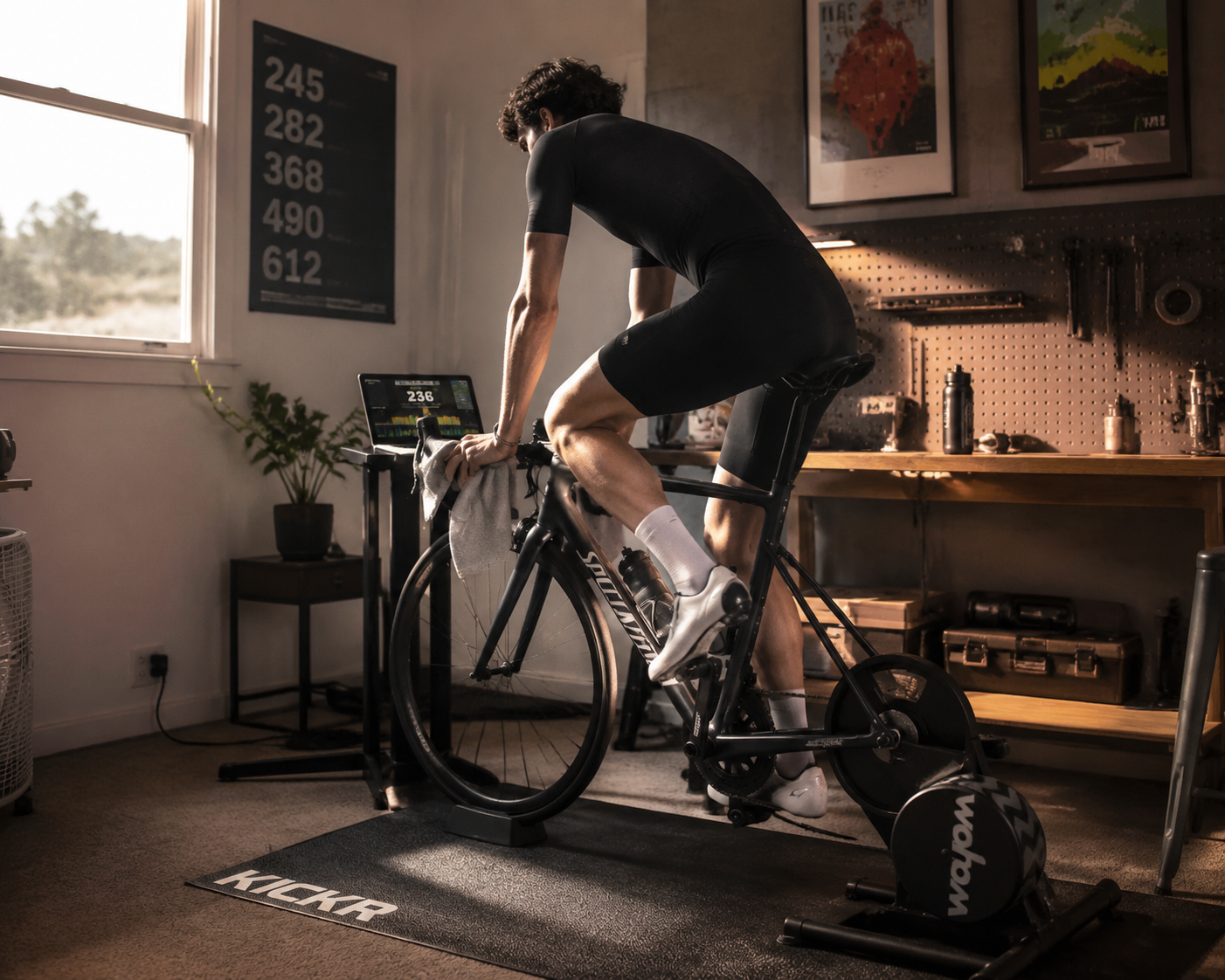 Home cyclist riding a smart trainer in an organized indoor training space with training data visible on a laptop