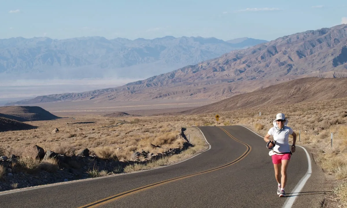 Runners on a long straight desert road through Death Valley with mountains in the distance and intense sunlight on the pavement
