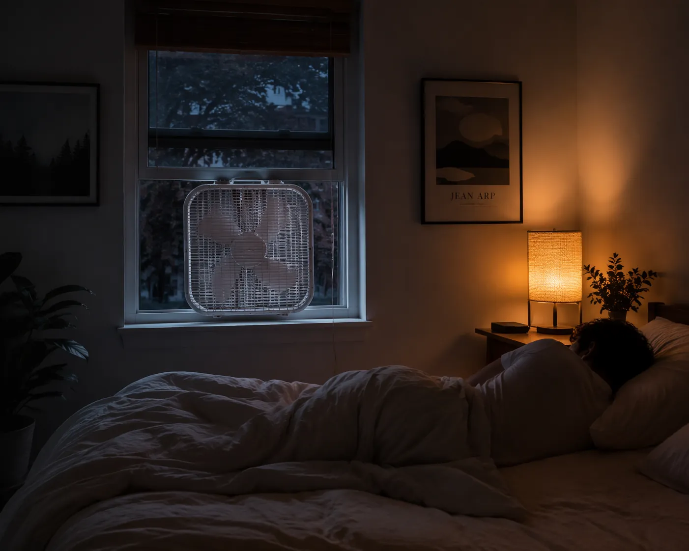 Warm bedroom at night arranged for sleeping without air conditioning with a box fan in the window and lightweight bedding